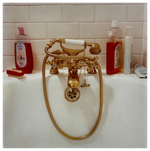 Load image into Gallery viewer, Photograph by Richard Heeps. Gold taps and hand held shower at the end of a bath. Red toned bottles of shampoo and baby bath contrast against the white bath and white bathroom tiles.
