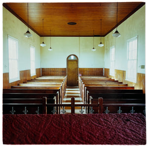 Photograph by Richard Heeps.  View of a small functional church from the purple pulpit cloth.  The pews are wooden and of basic design, the walls are painted white on the top half and wooden paneled on the bottom half.  Six basic pendular lights hang down from the wooden paneled ceiling.