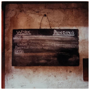 Photograph by Richard Heeps.  A chalk board with Work Pending written on the top with dates of 1976 and other chalk notes partially removed.  Chalk board hanging on a rough factory wall.
